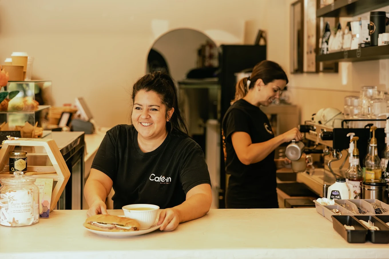 Deux membres de l'équipe du Café-in au comptoir : l'une sourit en servant un sandwich et une soupe, l'autre prépare un latté